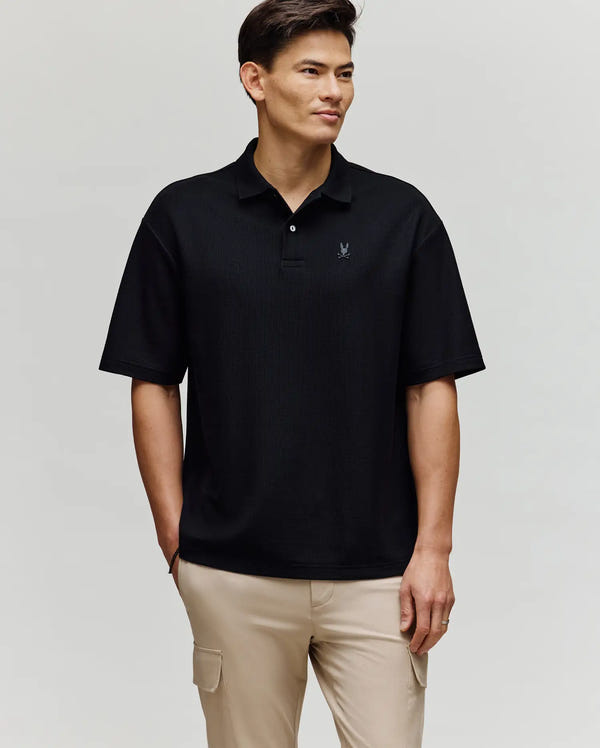 A man smiles and looks to the side, wearing a black Psycho Bunny MENS BENNY OVERSIZED PIQUE POLO SHIRT (MT0100105) in 100% Pima cotton with a chest logo, and beige cargo pants against a light background.