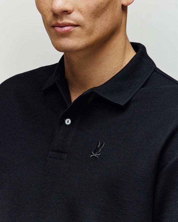 A person wears the Psycho Bunny MENS BENNY OVERSIZED PIQUE POLO SHIRT (MT0100105), black, 100% Pima cotton with an embroidered logo; only their lower face and upper torso are shown.