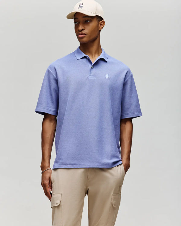 A man wearing the Psycho Bunny MENS BENNY OVERSIZED PIQUE POLO SHIRT (MT0100105) in light blue, paired with beige cargo pants and a beige cap, stands against a plain light gray background, looking slightly left with a neutral expression.