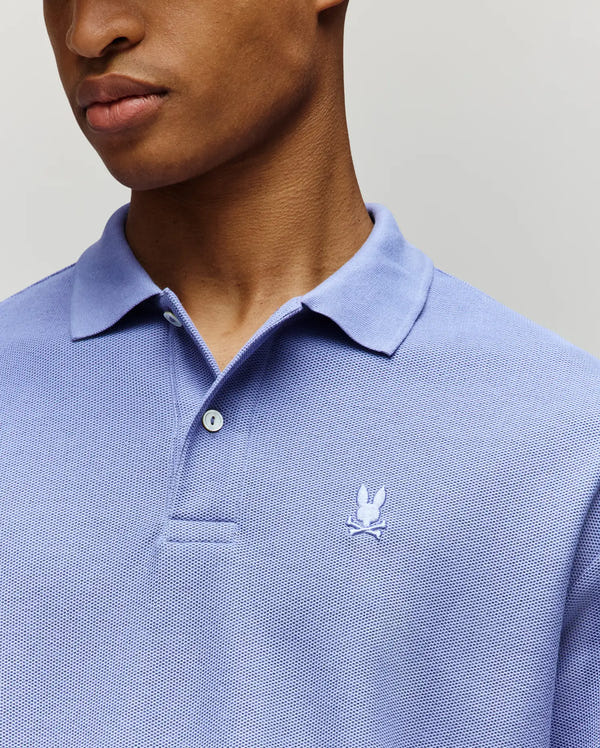 A person wears the Psycho Bunny MENS BENNY OVERSIZED PIQUE POLO SHIRT - MT0100105 in light blue, featuring a relaxed fit and a small white embroidered bunny logo on the left chest. The background is plain and the face is partially visible.