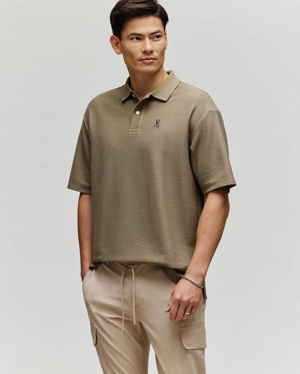 A man stands against a plain background wearing the Psycho Bunny MENS BENNY OVERSIZED PIQUE POLO SHIRT (MT0100105) in olive green with beige cargo pants. He has short dark hair, looks to the side, and has one hand in his pocket.