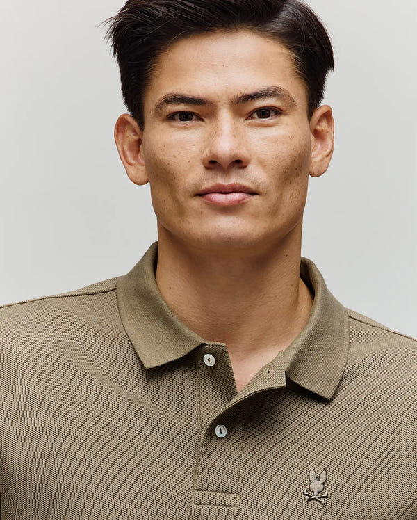 A man with short dark hair wears the Psycho Bunny MENS BENNY OVERSIZED PIQUE POLO SHIRT (MT0100105) in light brown, standing against a plain light background.