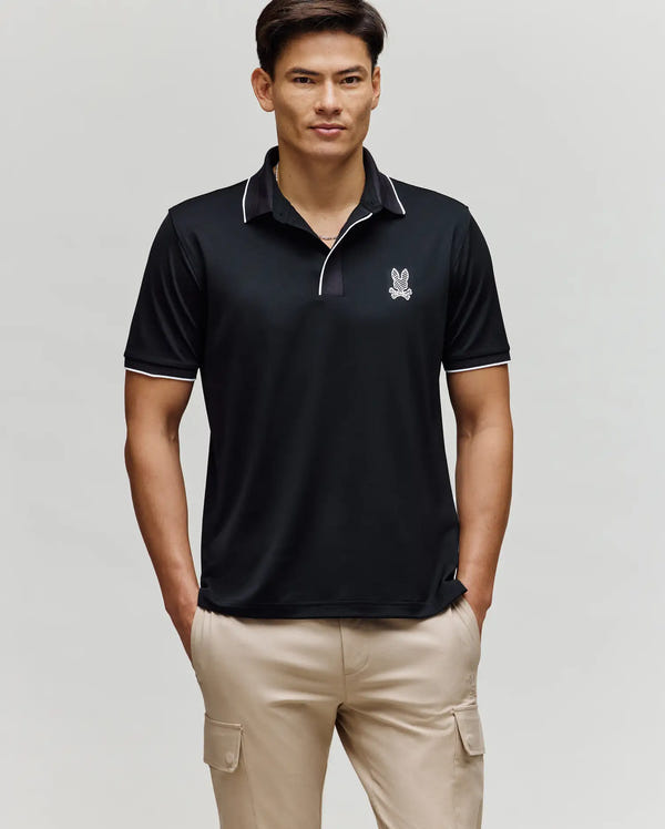 A man stands with his hands in his pockets, wearing the Psycho Bunny MENS JAYDEN SPORT POLO SHIRT - MT0100111 in black with white trim and a rabbit logo, paired with beige cargo pants, against a plain light background.
