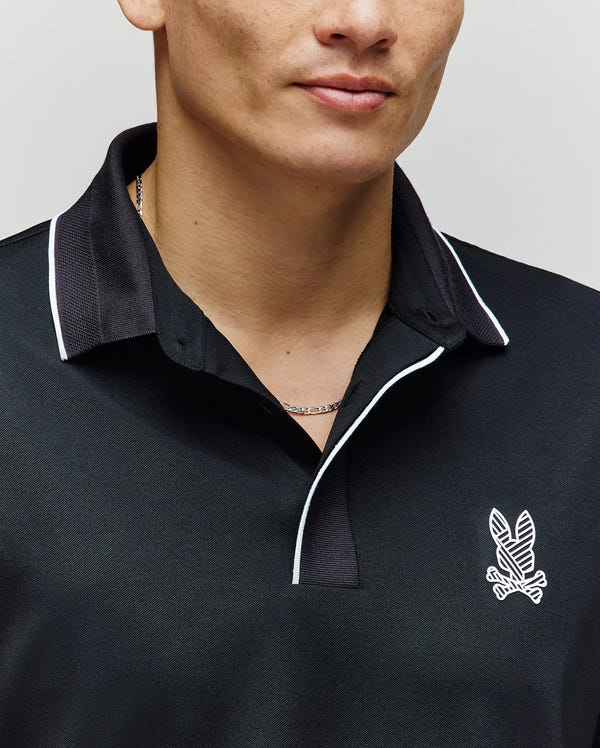 A man wears the Psycho Bunny MENS JAYDEN SPORT POLO SHIRT (MT0100111) in black with white trim and an embroidered winged logo. Only his lower face and upper torso are visible, highlighting the athletic fit and moisture-wicking fabric.