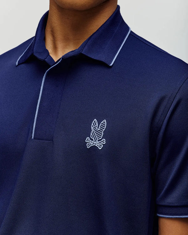 A man wears the Psycho Bunny MENS JAYDEN SPORT POLO SHIRT (MT0100111), navy blue with light blue collar trim and a white leaf with crossed bones logo on the chest. The image highlights its athletic fit, showing his upper torso and part of his face.