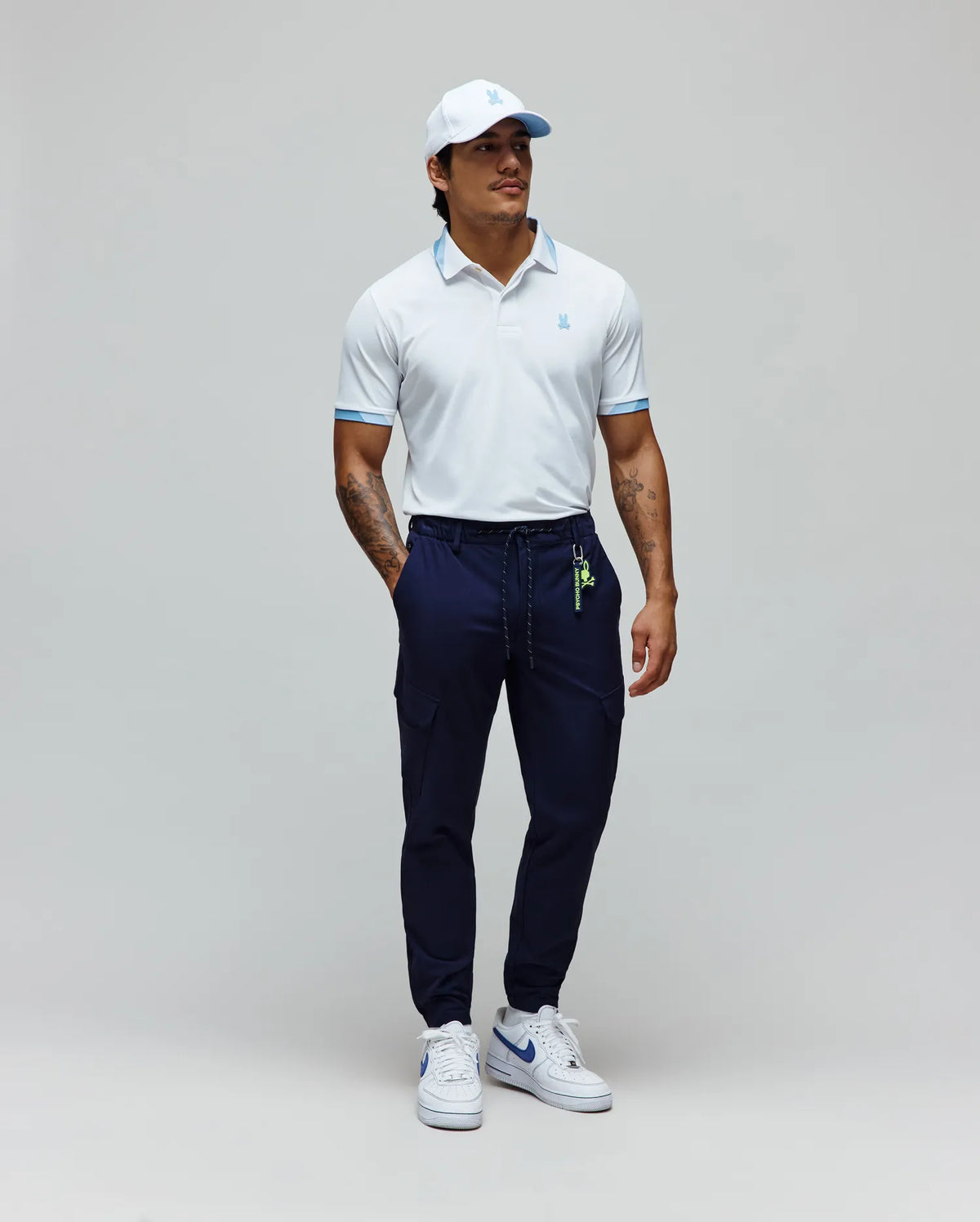 Mens Onyx Sport Polo, a white performance pique polo with light blue collar and sleeve accents, featuring a small embroidered Psycho Bunny skull logo on the chest, styled with navy cargo joggers and a white cap.