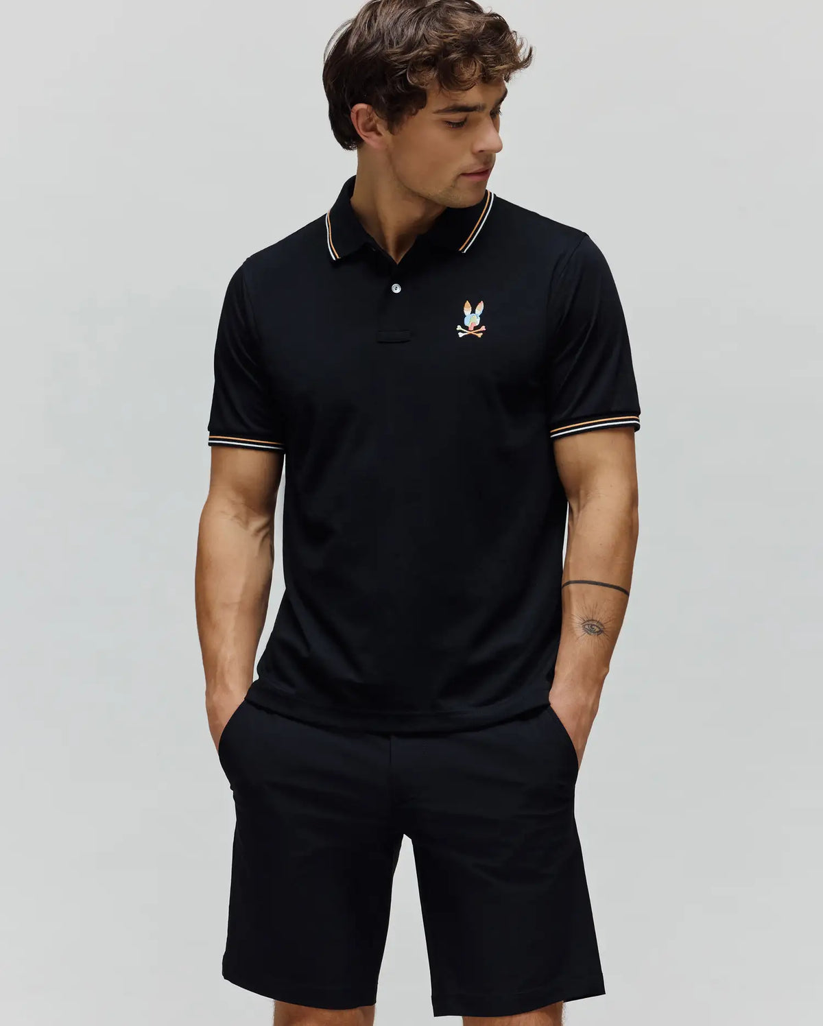 Black Mens Ara Pique Polo with a classic fit, featuring a pique texture, white and orange striped collar and cuffs, and a small, colorful embroidered Psycho Bunny skull logo on the left chest.