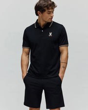 Black Mens Ara Pique Polo with a classic fit, featuring a pique texture, white and orange striped collar and cuffs, and a small, colorful embroidered Psycho Bunny skull logo on the left chest.