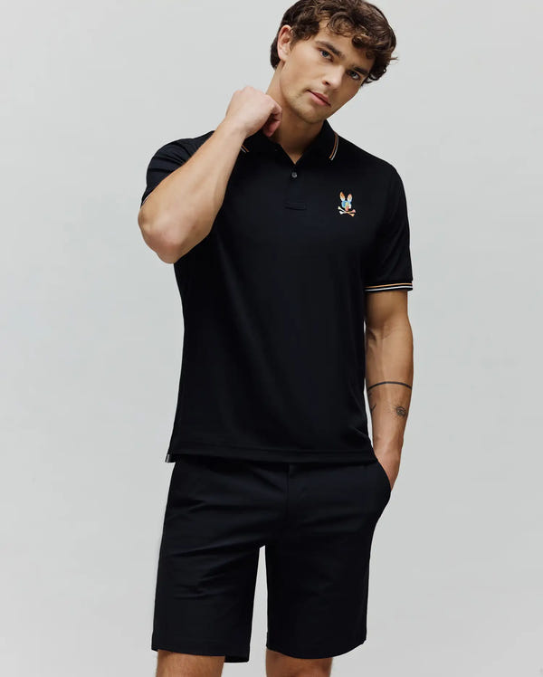 Mens Ara Pique Polo in black, featuring a classic fit, pique fabric, and a small, multi-colored embroidered Psycho Bunny skull logo on the left chest. Accent stripes in orange and white adorn the collar and sleeve cuffs.