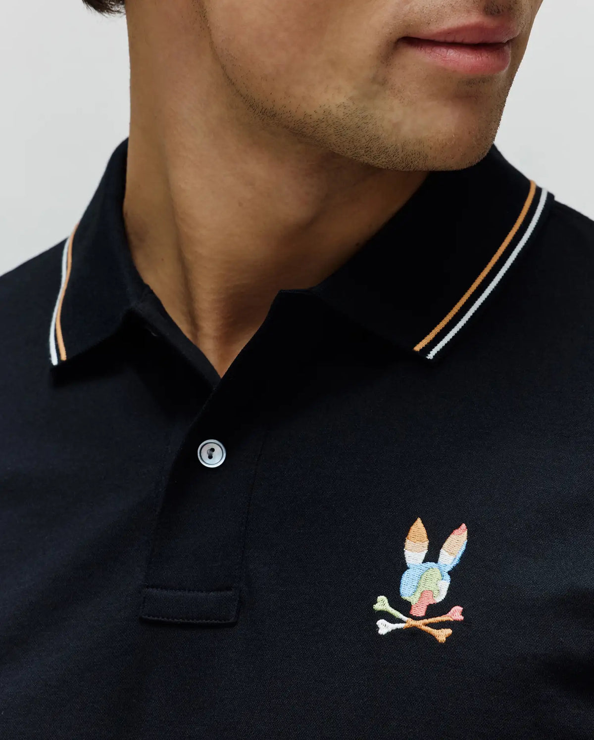 Black Mens Ara Pique Polo with classic fit, featuring a pique knit texture, white and orange striped collar, and a multicolored embroidered Psycho Bunny skull logo on the left chest.