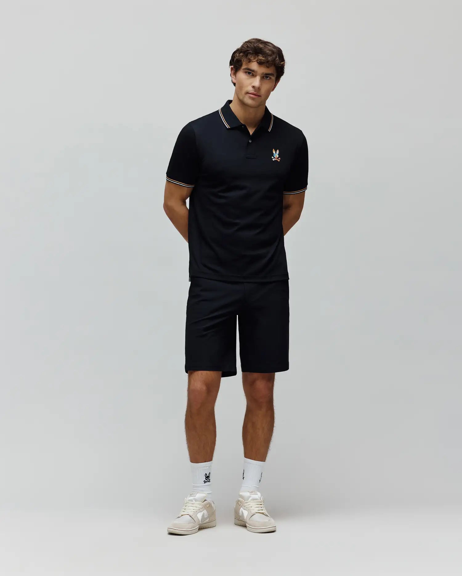 Mens Ara Pique Polo in navy, classic fit pique knit, with white and gold striped collar and cuffs, and a small embroidered multi-color Psycho Bunny skull logo on the left chest. Paired with matching navy shorts.