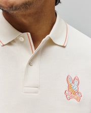 Mens Olivier Pique Polo in cream, classic fit, pique texture. Ribbed collar with orange stripe. Vibrant, multi-colored embroidered Psycho Bunny skull logo on left chest.