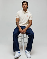 Mens Olivier Pique Polo in cream, a classic fit with pique texture, featuring orange tipping on the collar and cuffs. A vibrant orange embroidered Psycho Bunny skull logo adorns the left chest.