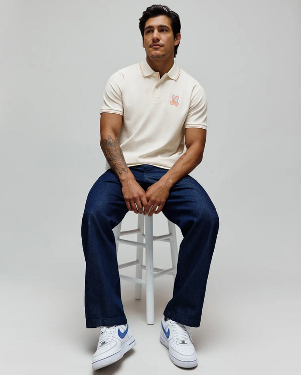 Mens Olivier Pique Polo in cream, a classic fit with pique texture, featuring orange tipping on the collar and cuffs. A vibrant orange embroidered Psycho Bunny skull logo adorns the left chest.