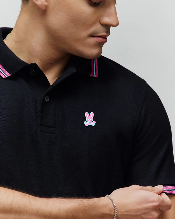 Mens Clark Pique Polo, a black classic fit pique polo shirt with vibrant pink and teal striped trim on the collar and sleeve cuffs, featuring a small embroidered pink and light blue Psycho Bunny skull logo on the left chest.