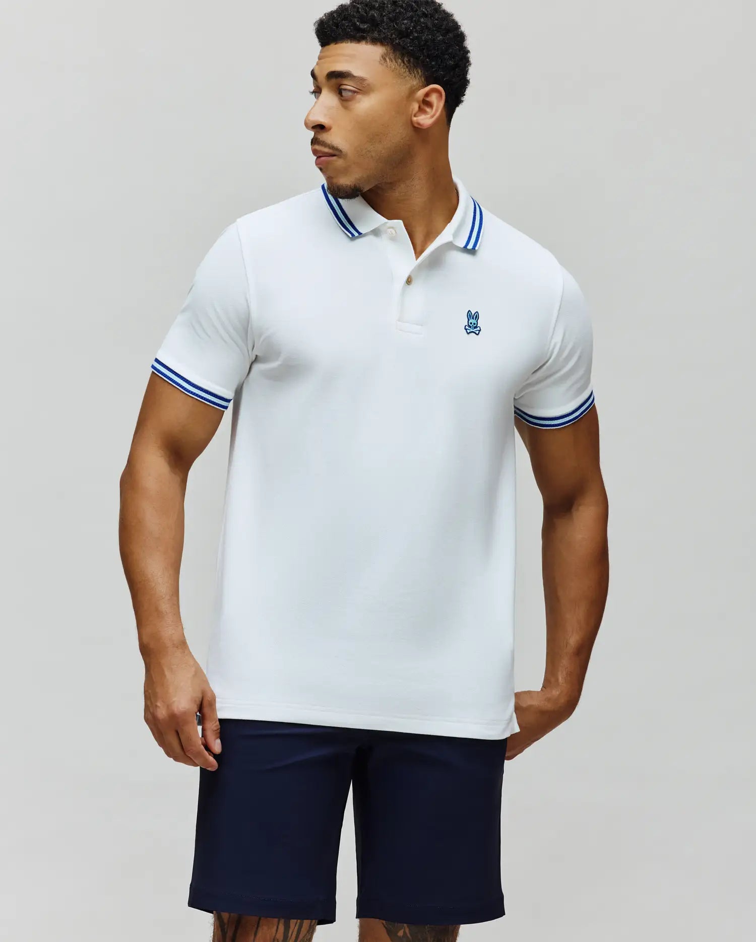 Mens Clark Pique Polo, a classic fit white pique polo shirt with a blue embroidered Psycho Bunny skull logo on the left chest and double blue tipping on the collar and sleeve cuffs.