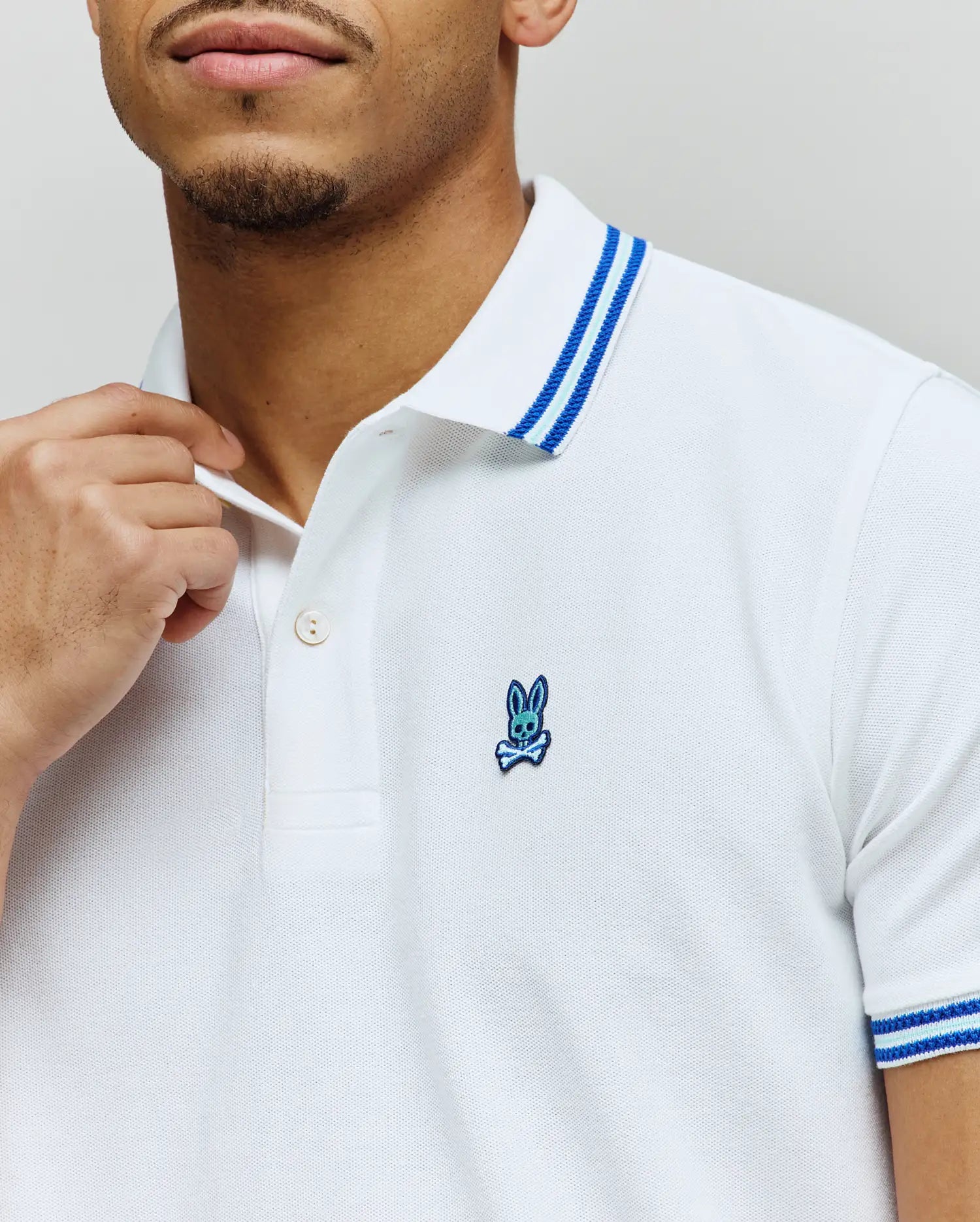 White Mens Clark Pique Polo, classic fit, featuring textured pique fabric, a small embroidered blue Psycho Bunny skull logo on the left chest, and blue striped collar and sleeve cuffs.