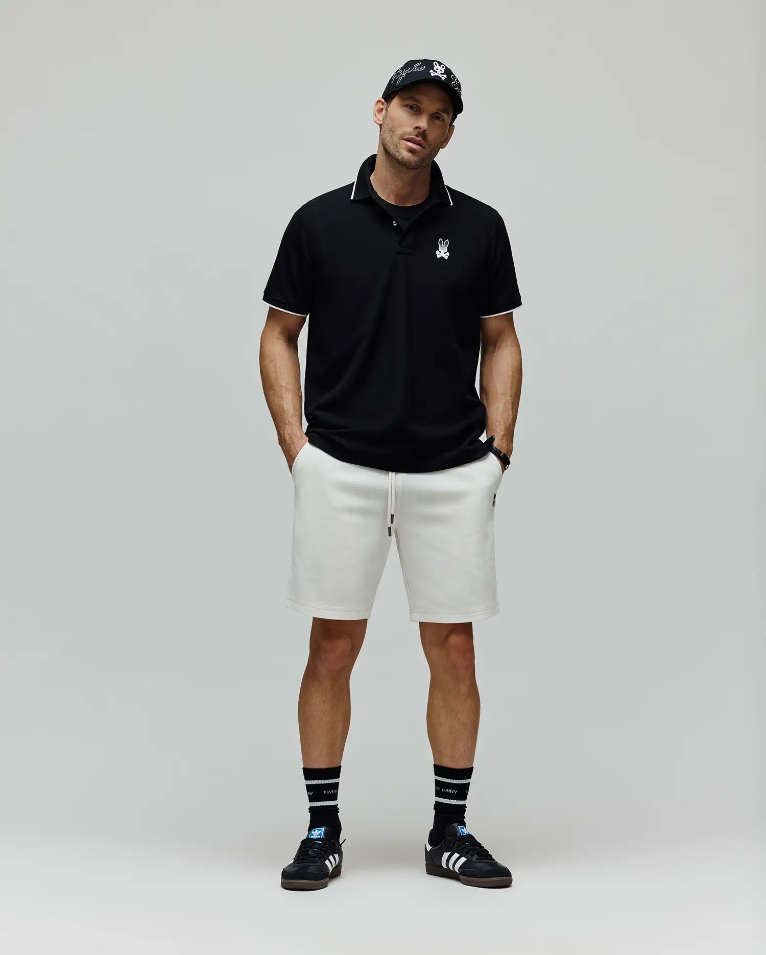 Mens Aram Pique Polo, a black classic fit pique polo with white tipping on collar and cuffs, featuring a small white embroidered Psycho Bunny skull logo on the left chest. Paired with ecru relaxed French terry shorts with a small black embroidered logo.