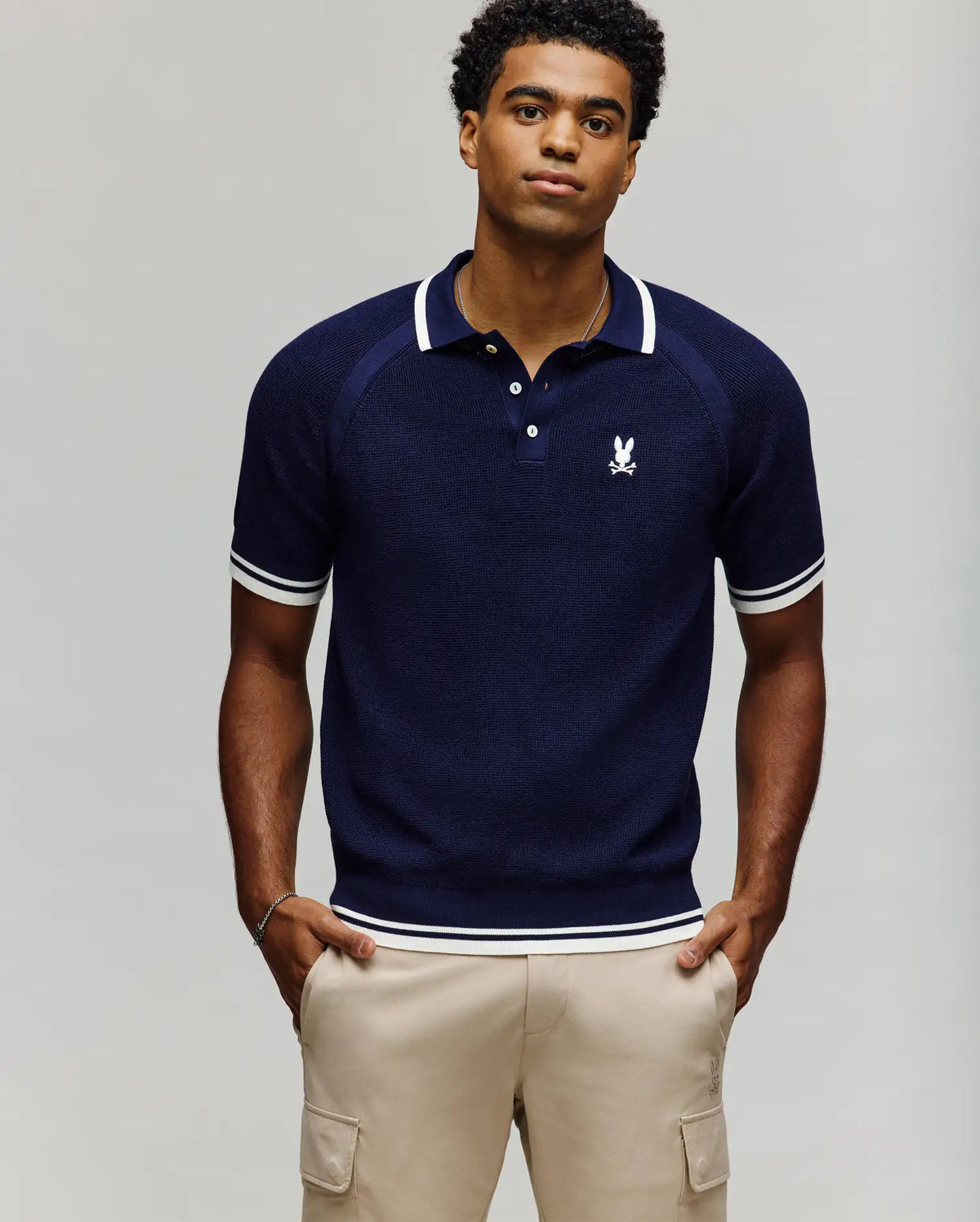 A man with short curly hair stands against a plain background, hands in pockets, wearing Psycho Bunny’s MENS LAZARE KNIT POLO - MT0100165 in navy with white trim and embroidery, paired with beige cargo pants.