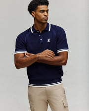 A man stands with arms crossed, looking to the side, wearing the Psycho Bunny MENS LAZARE KNIT POLO - MT0100165 in navy blue with white trim and beige pants against a plain light background.