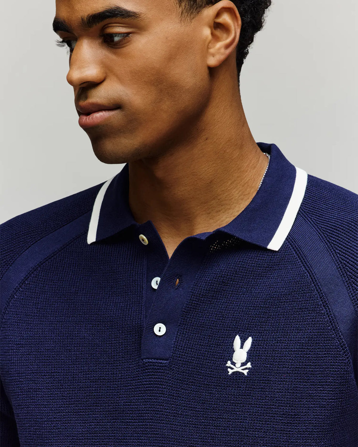 A man wearing the Psycho Bunny MENS LAZARE KNIT POLO - MT0100165, a navy blue colorblock cotton polo with white trim and an embroidered bunny and crossbones logo on the chest, looks to his left against a neutral background.