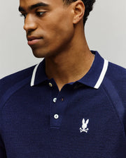A man wearing the Psycho Bunny MENS LAZARE KNIT POLO - MT0100165, a navy blue colorblock cotton polo with white trim and an embroidered bunny and crossbones logo on the chest, looks to his left against a neutral background.