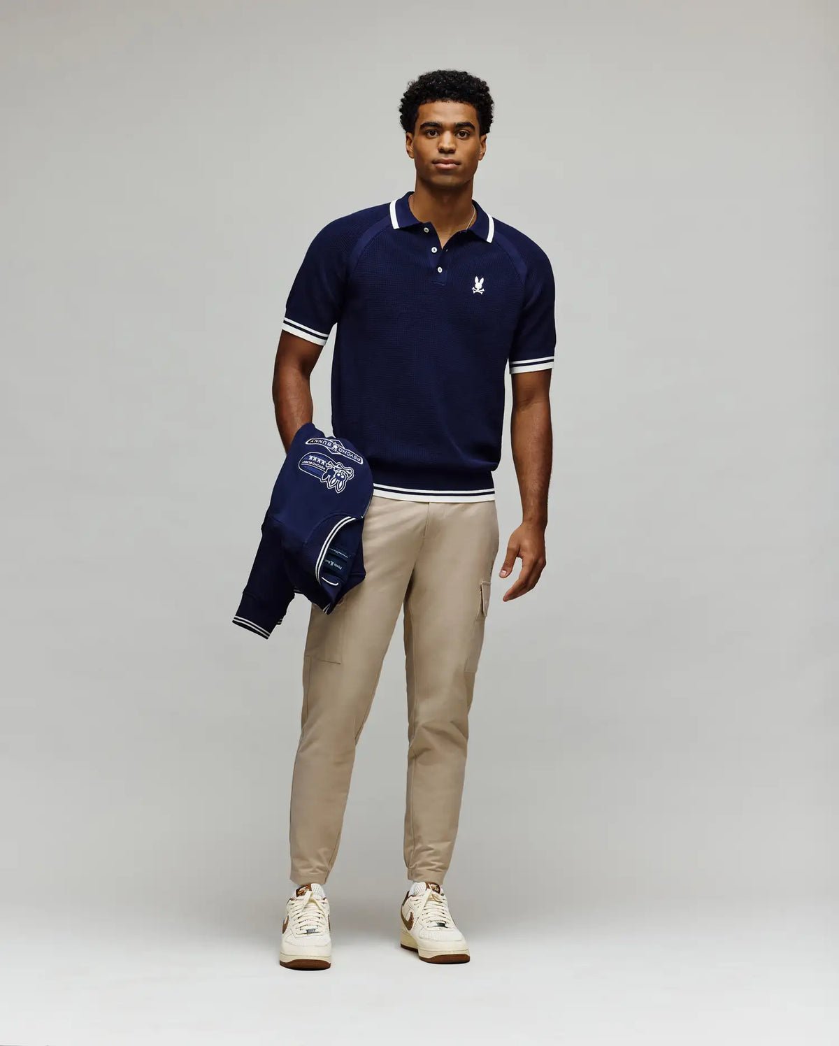 A man faces forward against a light gray background, wearing the Psycho Bunny MENS LAZARE KNIT POLO (MT0100165) in navy, beige pants, and white sneakers, holding a navy jacket in his left hand.