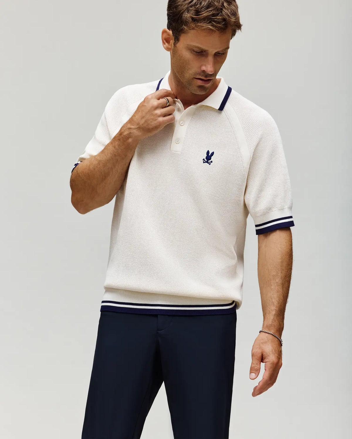 A man wears the Psycho Bunny MENS LAZARE KNIT POLO (MT0100165), a short-sleeve cream textured polo with navy trim and a small navy emblem on the chest, paired with dark pants, standing against a plain light background.
