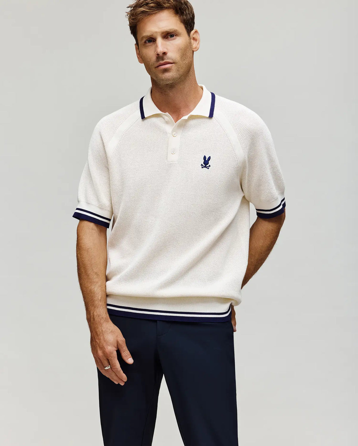 A man stands against a plain light background wearing the MENS LAZARE KNIT POLO (MT0100165) by Psycho Bunny—a cream, short-sleeve colorblock polo with navy trim and an embroidered bunny logo on the chest—paired with dark pants.