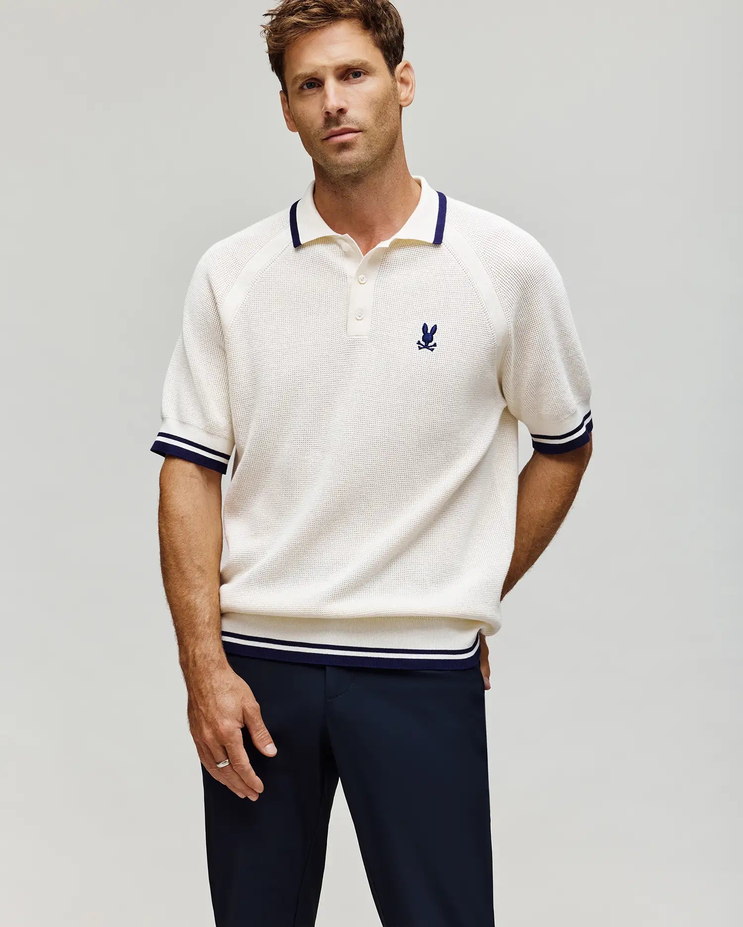 A man stands against a plain light background wearing the MENS LAZARE KNIT POLO (MT0100165) by Psycho Bunny—a cream, short-sleeve colorblock polo with navy trim and an embroidered bunny logo on the chest—paired with dark pants.