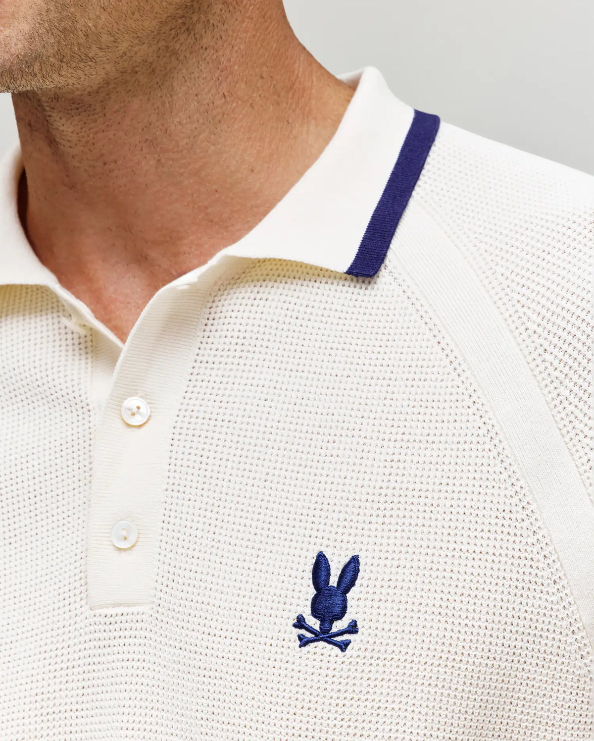 Close-up of a person wearing the Psycho Bunny MENS LAZARE KNIT POLO (MT0100165), featuring cream-colored textured knit, blue collar stripe, and embroidered bunny logo above crossed bones. Only the lower face and upper chest are visible.