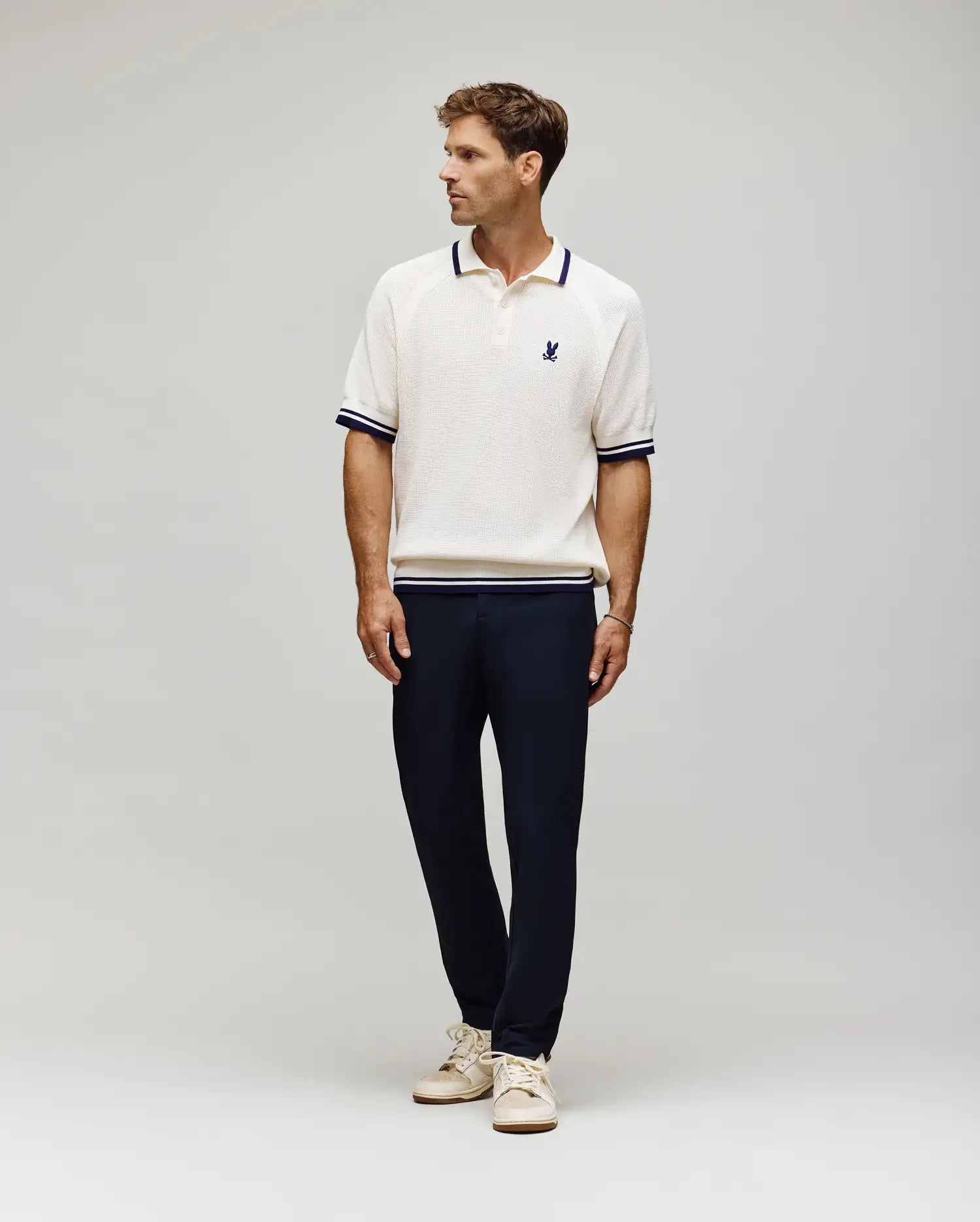 A man stands against a plain background wearing the Psycho Bunny MENS LAZARE KNIT POLO (MT0100165) in white with navy trim, paired with navy pants and beige sneakers. He looks to the side, hands relaxed at his sides.
