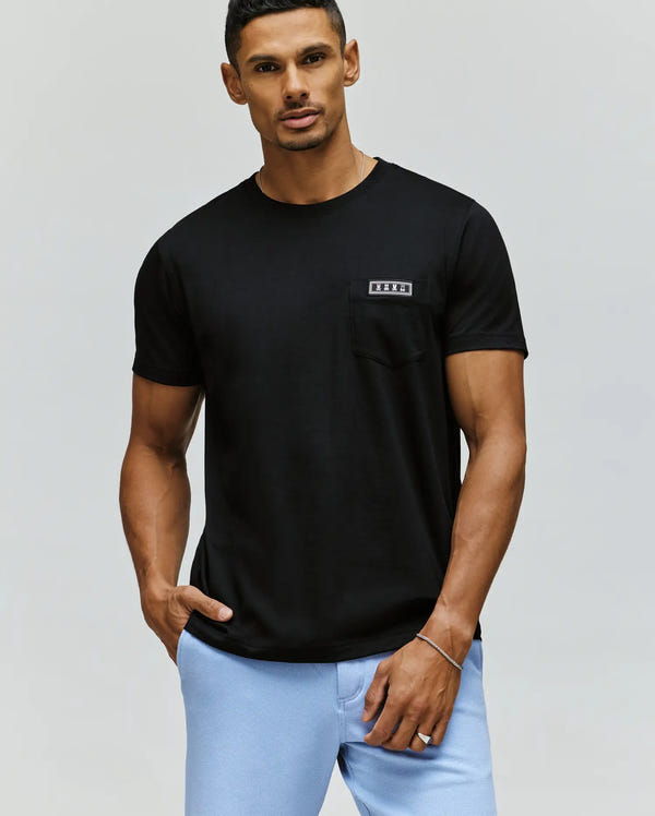 A man stands with one hand in his pocket against a plain light background, wearing light blue pants and a black Psycho Bunny MENS AXTON POCKET TEE (MT0200001) made from Pima cotton, featuring a small chest pocket.