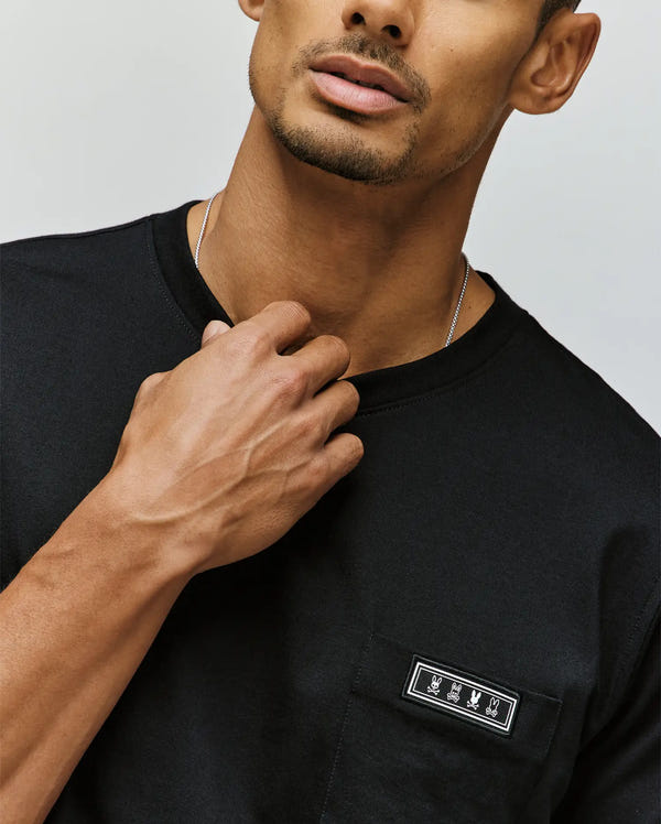 A man wears the Psycho Bunny MENS AXTON POCKET TEE (MT0200001), a black Pima cotton shirt with a small white hand symbol patch on the pocket, holding the neckline. His face is partly visible, displaying a trimmed beard.