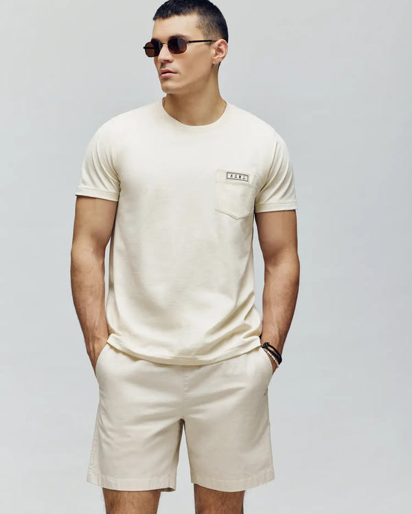 A man wearing sunglasses, a Psycho Bunny Mens Axton Pocket Tee (MT0200001) in light beige Pima cotton with a chest pocket, and matching beige shorts stands with his hands in his pockets against a plain background.