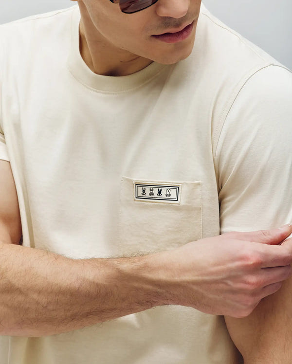A man in sunglasses, looking down and adjusting his sleeve, wears the Psycho Bunny MENS AXTON POCKET TEE - MT0200001 in beige, made from Pima cotton with a pocket featuring three black embroidered bunnies.