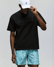 A person in a white bunny-logo cap, turquoise shorts with a blue bunny pattern, and the Psycho Bunny MENS ZAYN OVERSIZED GRAPHIC TEE (MT0200004) in black stands against a plain light background.