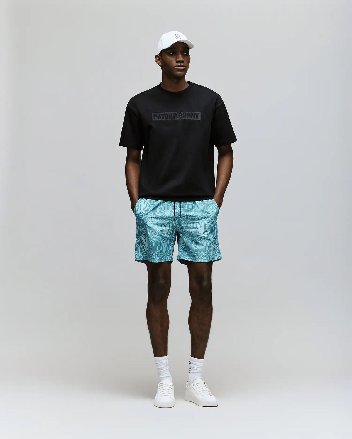 A man wears the Psycho Bunny MENS ZAYN OVERSIZED GRAPHIC TEE (MT0200004) in black, blue patterned shorts, white socks and sneakers, and a white cap, standing facing forward with hands in pockets against a plain background.