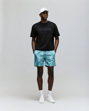 A man wears the Psycho Bunny MENS ZAYN OVERSIZED GRAPHIC TEE (MT0200004) in black, blue patterned shorts, white socks and sneakers, and a white cap, standing facing forward with hands in pockets against a plain background.