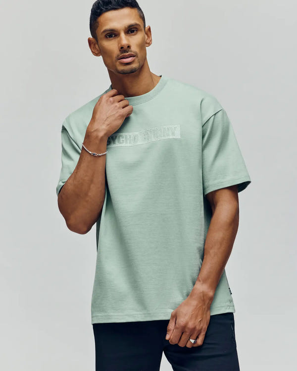 A man stands against a plain background wearing the Psycho Bunny MENS ZAYN OVERSIZED GRAPHIC TEE (MT0200004), a light green heavyweight jersey shirt with "PSYCHO BUNNY" faintly printed across the chest, one hand touching his neck.