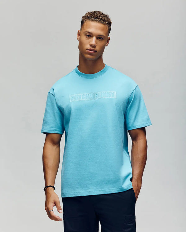 A young man with short curly hair wears a relaxed fit Psycho Bunny MENS ZAYN OVERSIZED GRAPHIC TEE - MT0200004 in light blue with subtle white chest text and black pants, standing against a plain light gray background.