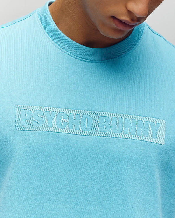 A person wears the Psycho Bunny MENS ZAYN OVERSIZED GRAPHIC TEE (MT0200004), featuring bold lettering across the chest. The relaxed, oversized fit gives a casual vibe, with only their lower face and neck partially visible.