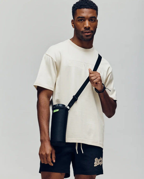 A man stands against a plain background, wearing the Psycho Bunny MENS ZAYN OVERSIZED GRAPHIC TEE (MT0200004) in cream and black shorts. He carries a black water bottle in a shoulder strap holder, gazing forward with a neutral expression.