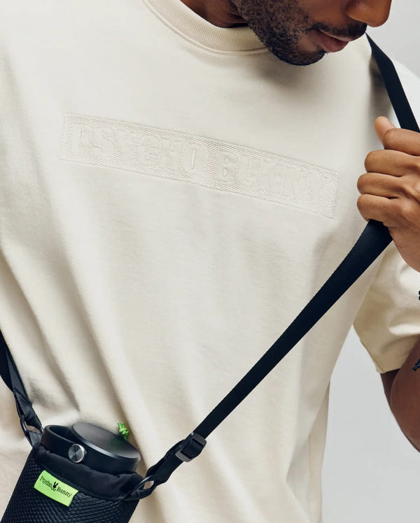 A person in a comfy, off-white Psycho Bunny MENS ZAYN OVERSIZED GRAPHIC TEE (MT0200004) holds the strap of a crossbody bag with a green tag. Only the lower half of their face is shown.