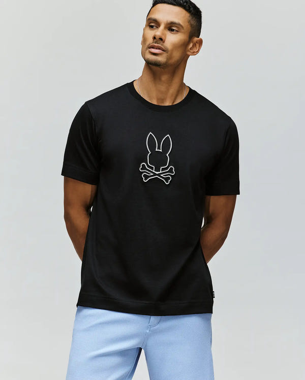 A man stands with his hands behind his back, looking to the side, wearing the Psycho Bunny MENS SETH GRAPHIC TEE (MT0200005) in 100% Pima cotton with a white bunny and crossbones graphic, paired with light blue shorts.