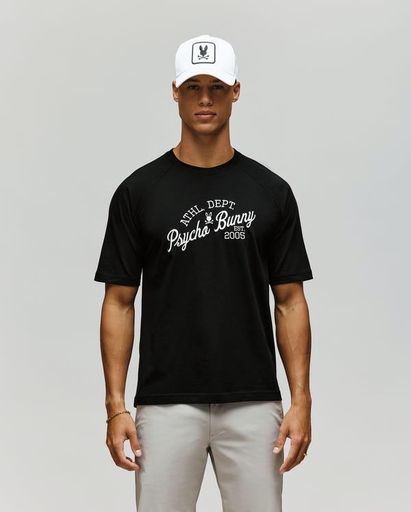 A man stands facing forward against a plain light background, wearing a white cap, light gray pants, and the Psycho Bunny MENS MATTEO RAGLAN GRAPHIC TEE (MT0200008), a black t-shirt with "PHL DEPT Psycho Bunny 2005" on it.