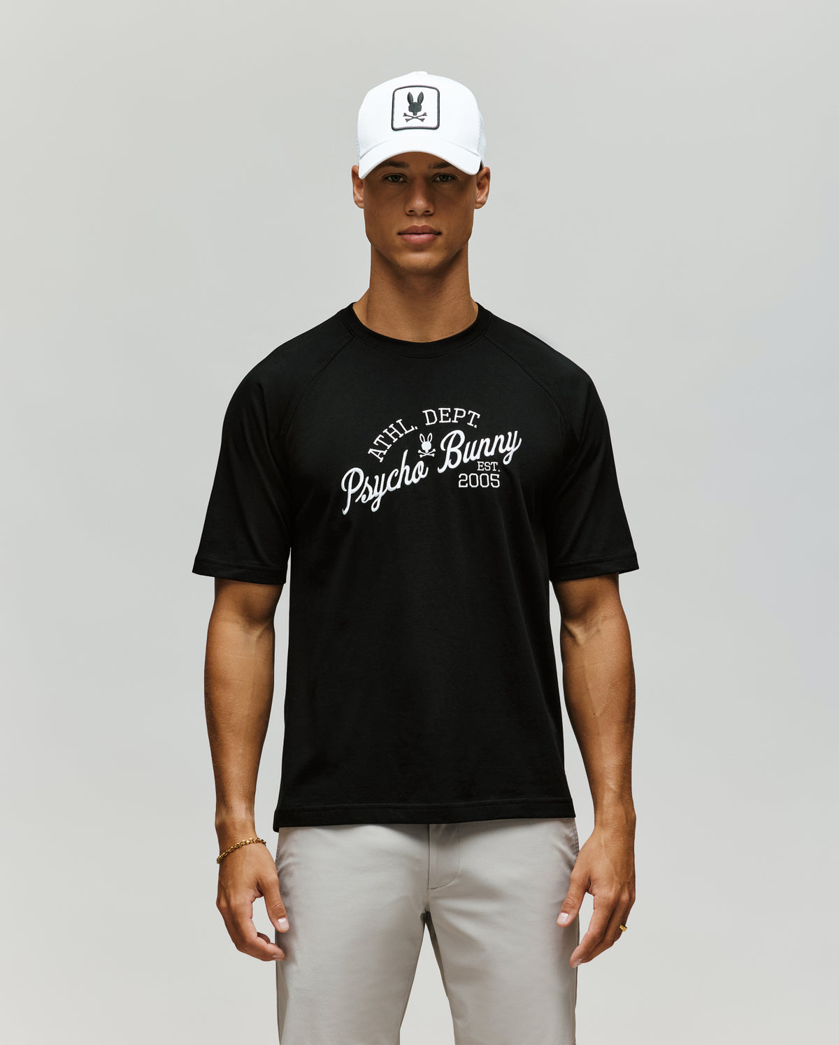 A man stands facing forward against a plain light background, wearing a white cap, light gray pants, and the Psycho Bunny MENS MATTEO RAGLAN GRAPHIC TEE (MT0200008), a black t-shirt with "PHL DEPT Psycho Bunny 2005" on it.