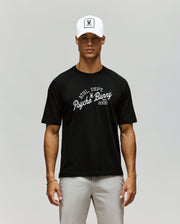 A man stands facing forward against a plain light background, wearing a white cap, light gray pants, and the Psycho Bunny MENS MATTEO RAGLAN GRAPHIC TEE (MT0200008), a black t-shirt with "PHL DEPT Psycho Bunny 2005" on it.