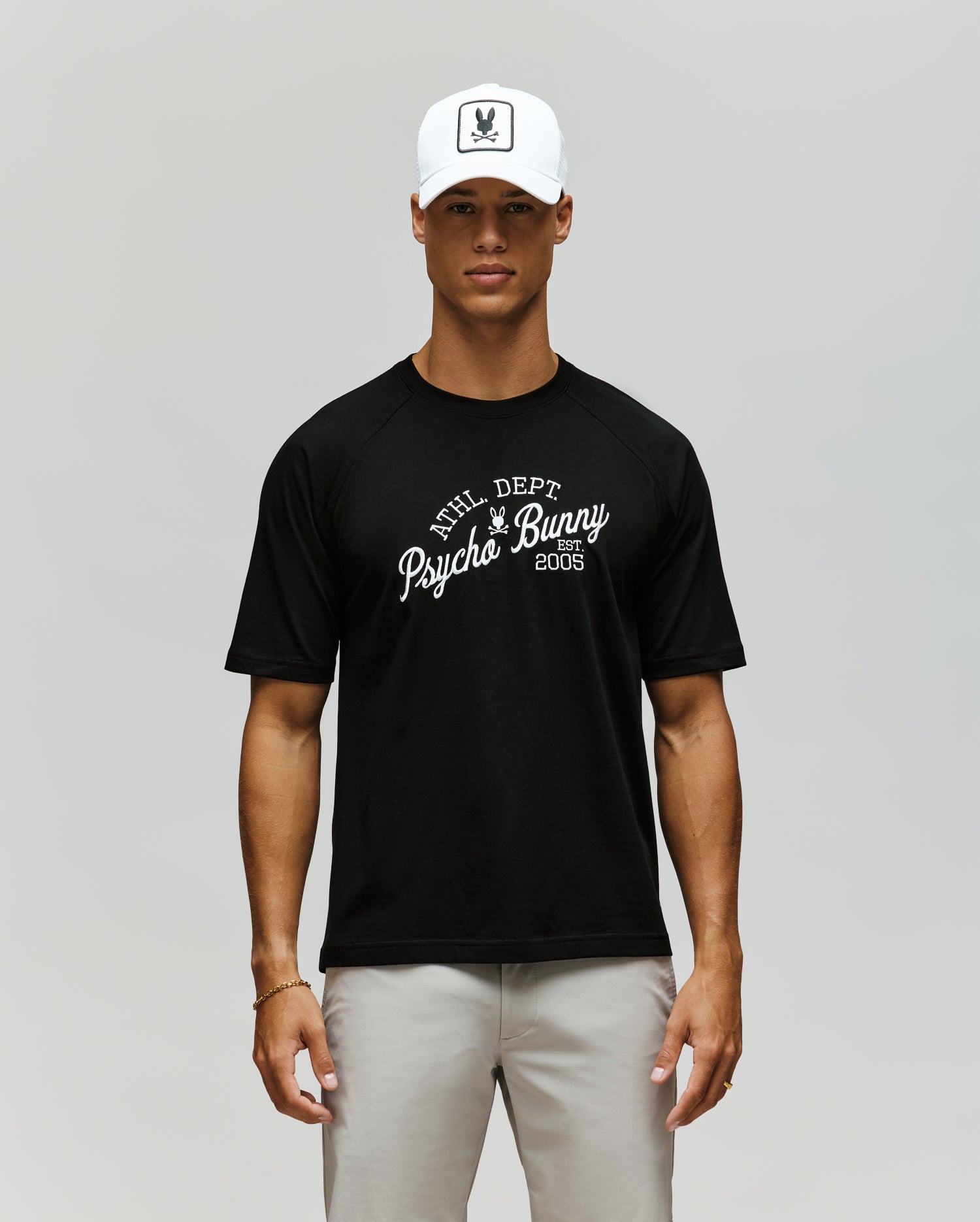 A man stands facing forward against a plain light background, wearing a white cap, light gray pants, and the Psycho Bunny MENS MATTEO RAGLAN GRAPHIC TEE (MT0200008), a black t-shirt with "PHL DEPT Psycho Bunny 2005" on it.