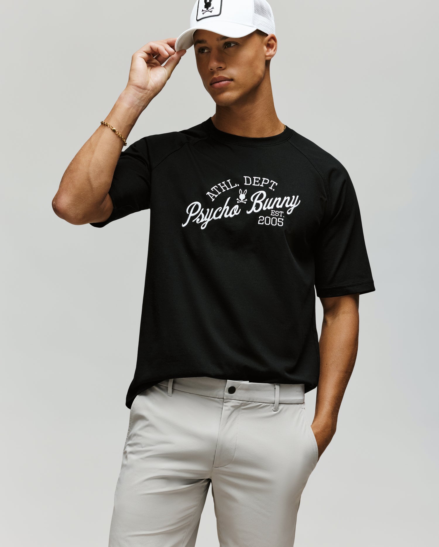 A man in light gray pants and a white cap stands against a plain background, wearing the Psycho Bunny MENS MATTEO RAGLAN GRAPHIC TEE (MT0200008) with "Psycho Bunny" and "2005" printed on his black t-shirt, holding the brim of his cap.